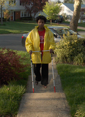 Photo shows Kelly walking along the sidewalk from her driveway with a rectangular AMD that looks like two white canes with crossbars.  The bottom of the AMD has stopped at the bottom of a step.